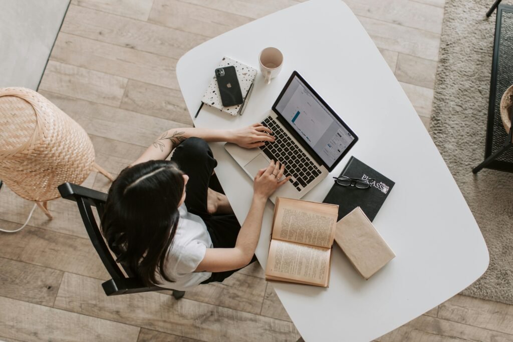 Overhead view of a woman using a laptop while searching for the Ericksonian hypnosis glossary at a home desk, surrounded by books, a phone, and a cup.