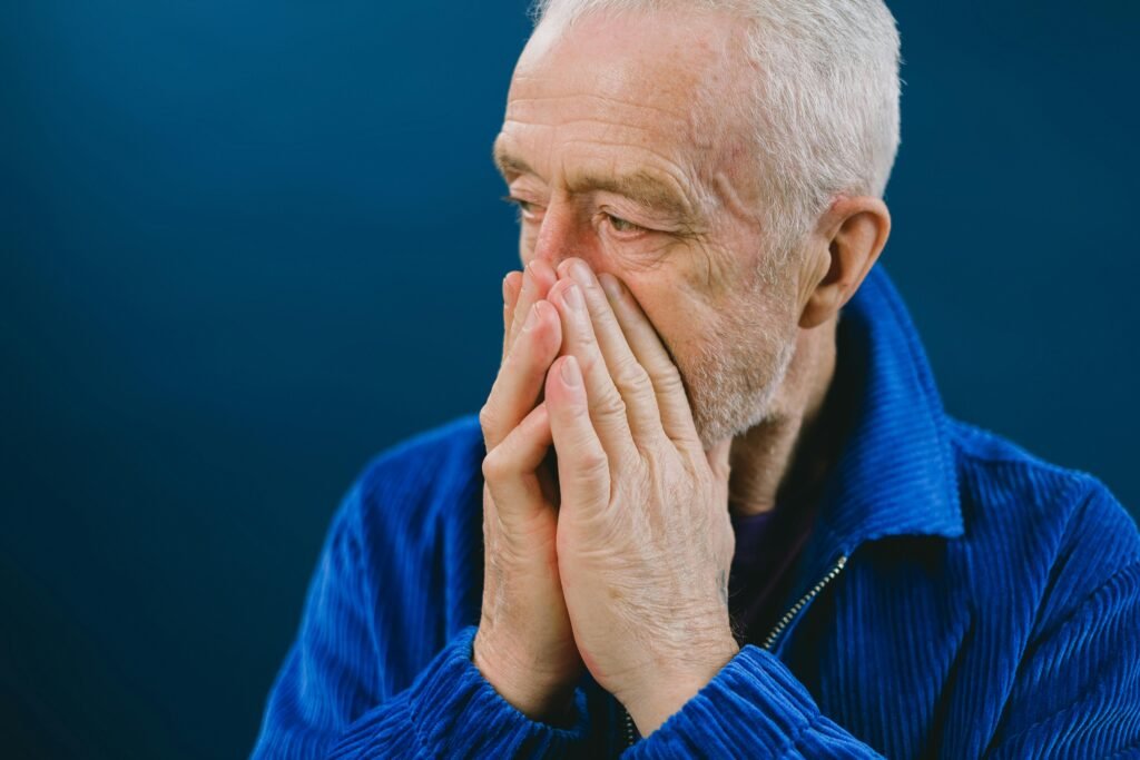 Serious anxious bearded senior gray haired male in blue jacket covering mouth with hands while looking away against blue background